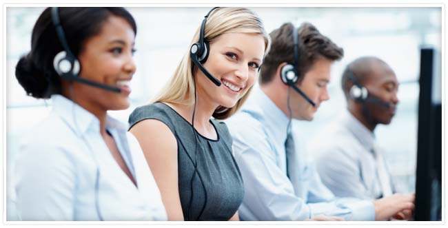 Four customer support employees wearing headsets sitting next to each other with one blonde employee smiling and looking at you.