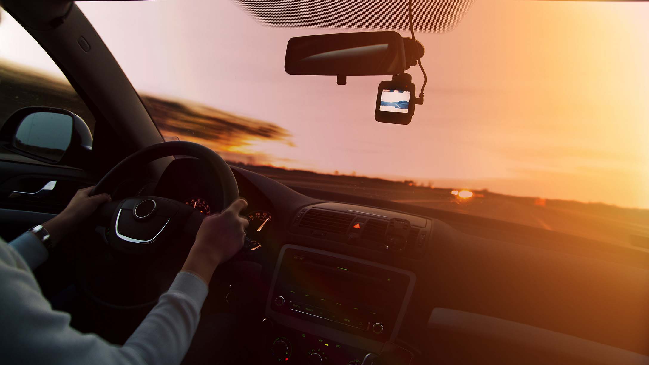 Vista desde el interior de un vehículo en movimiento, con el conductor al volante y una cámara de tablero instalada en el parabrisas, una solución de cámaras para vehículos. El sol se está poniendo en el horizonte, creando un resplandor cálido.