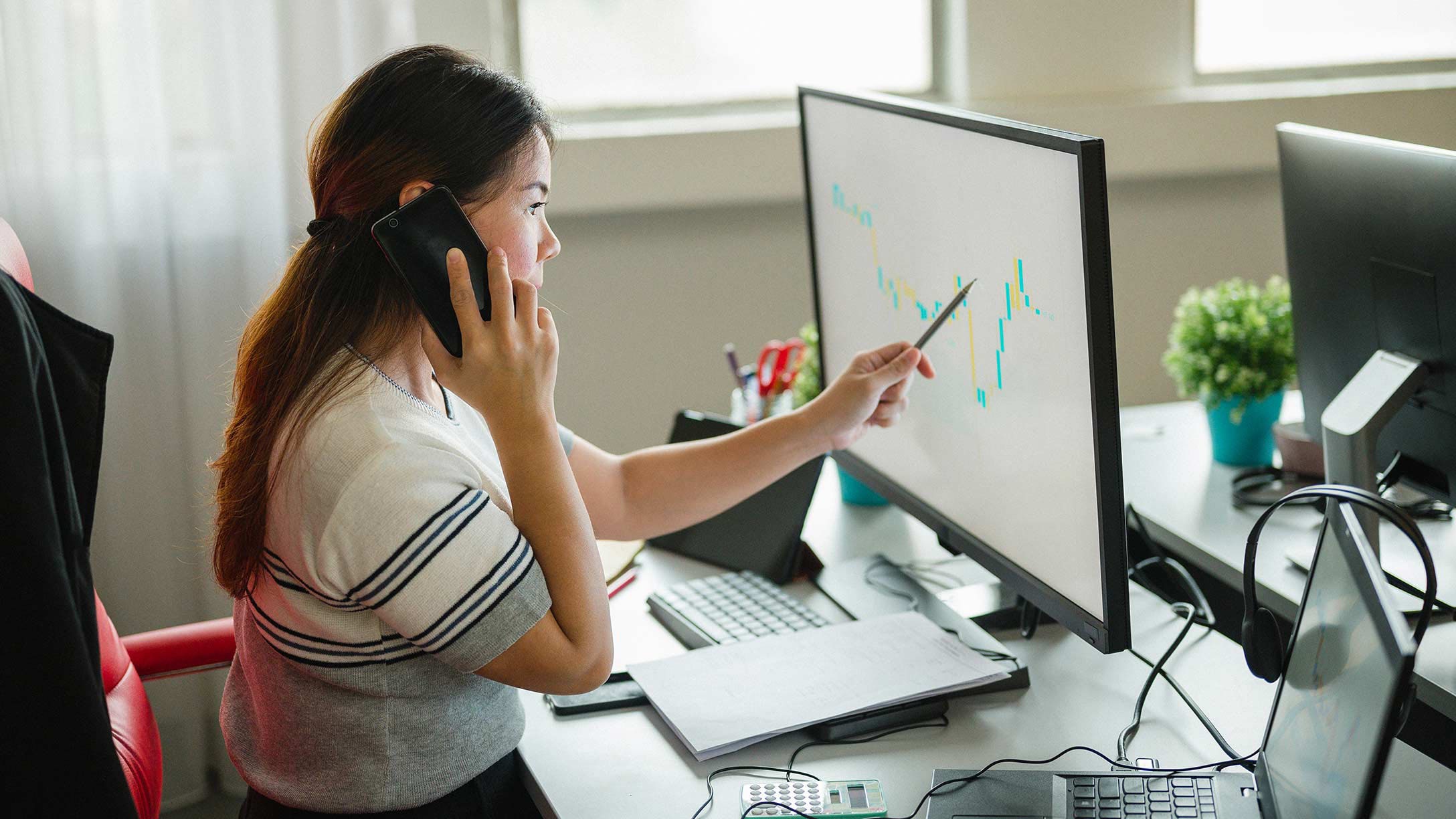 Una donna seduta davanti al computer con il telefono all'orecchio e una penna che punta allo schermo