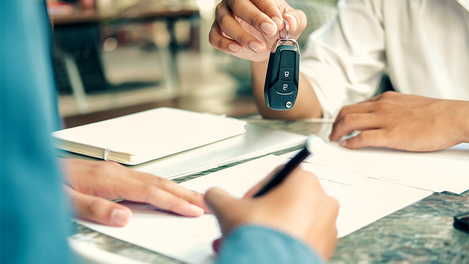 Foto di una scrivania e una persona che firma documenti e dall'altra parte una mano tende un mazzo di chiavi di un'automobile