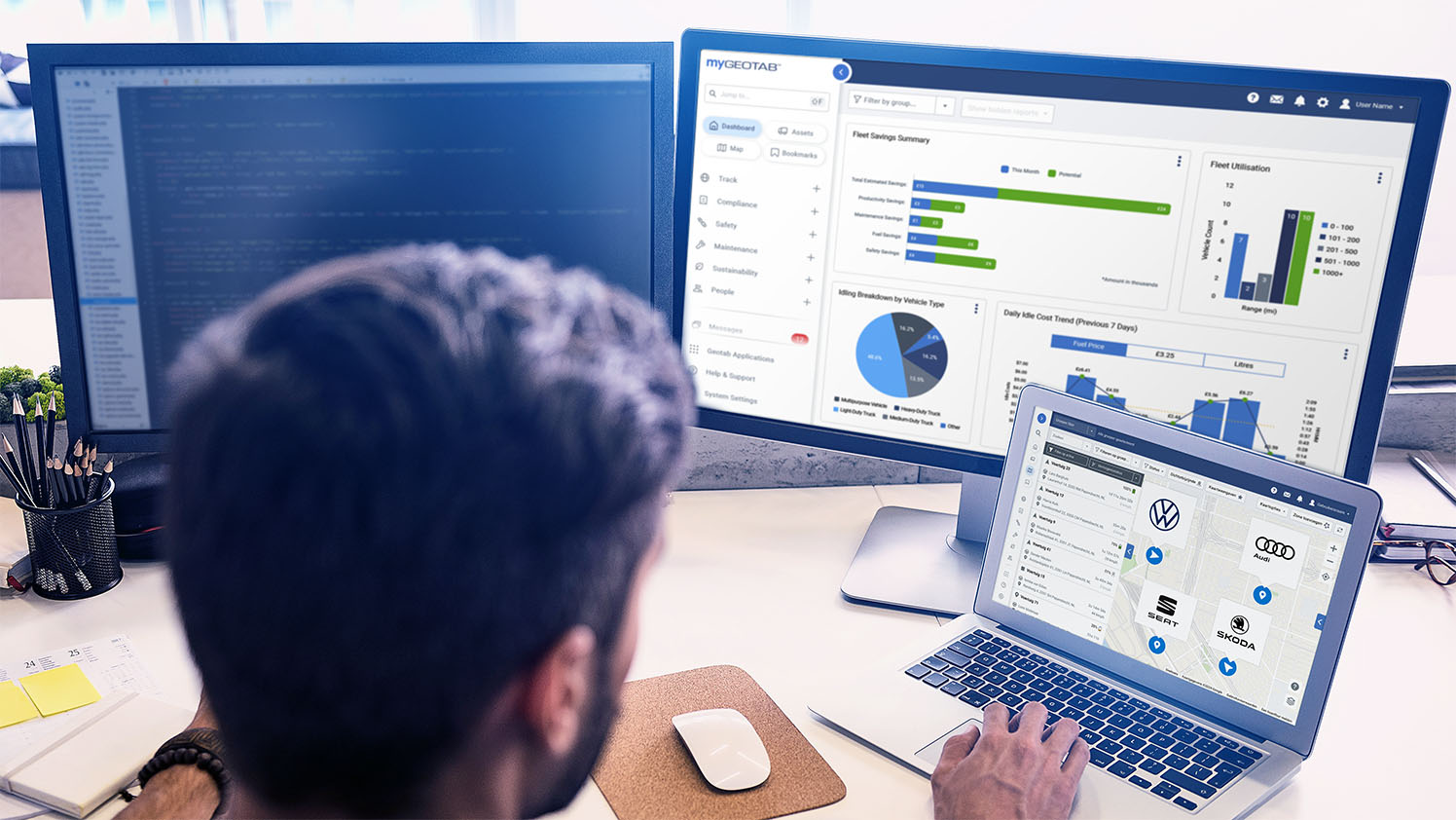 person at a desk with a laptop and a monitor showing MyGeotab UI